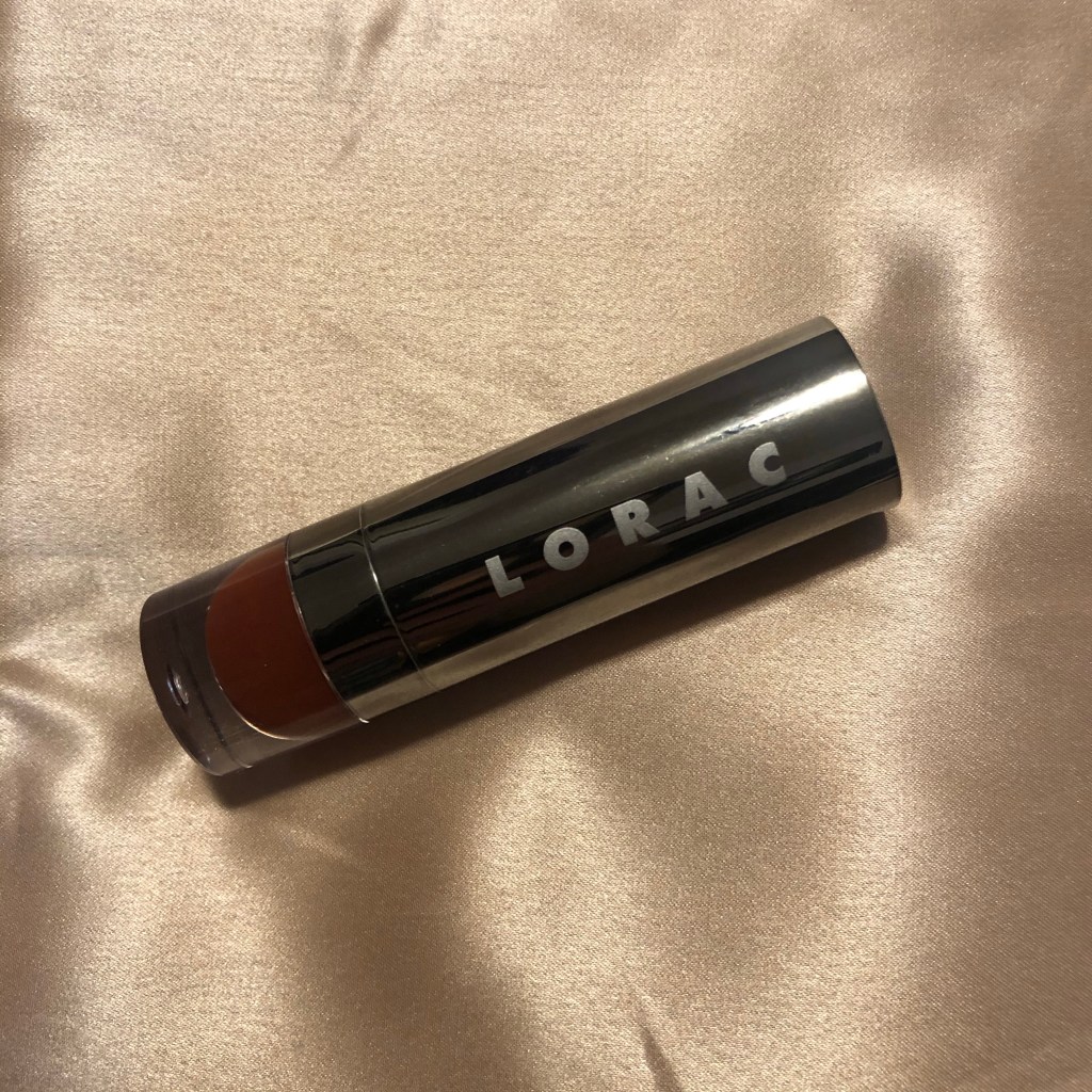 A photo of the Lorac Alter Ego Lipstick in shade Hipster. The color is a rusted deep red brown and the tube rests on a caramel silk sheet. 