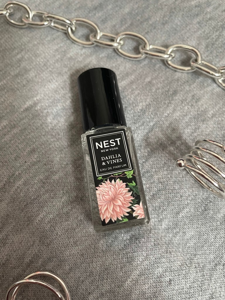 A picture of the travel size fragrance Dahlia and Vines from NEST New York Fragrances on a gray background surrounded by silver jewelry.