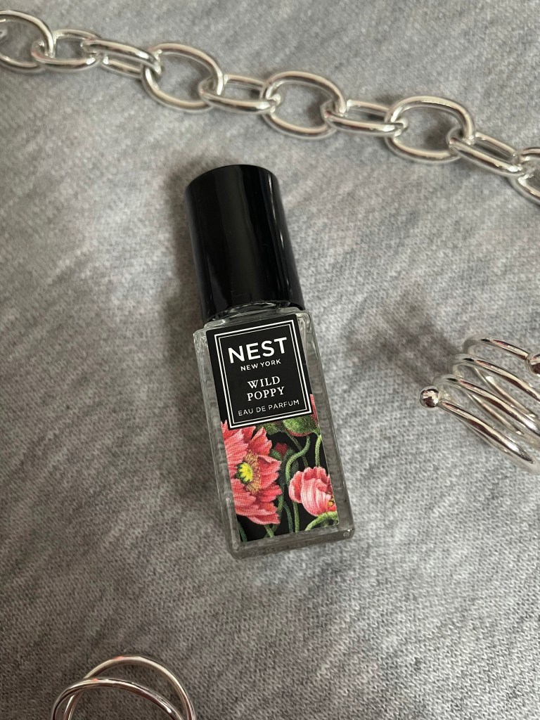 A picture of the travel size fragrance Wild Poppy from NEST New York Fragrances on a gray background surrounded by silver jewelry.
