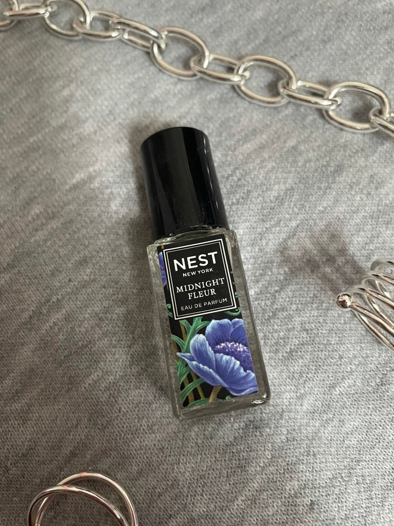 A picture of the travel size fragrance Midnight Fleur from NEST New York Fragrances on a gray background surrounded by silver jewelry.