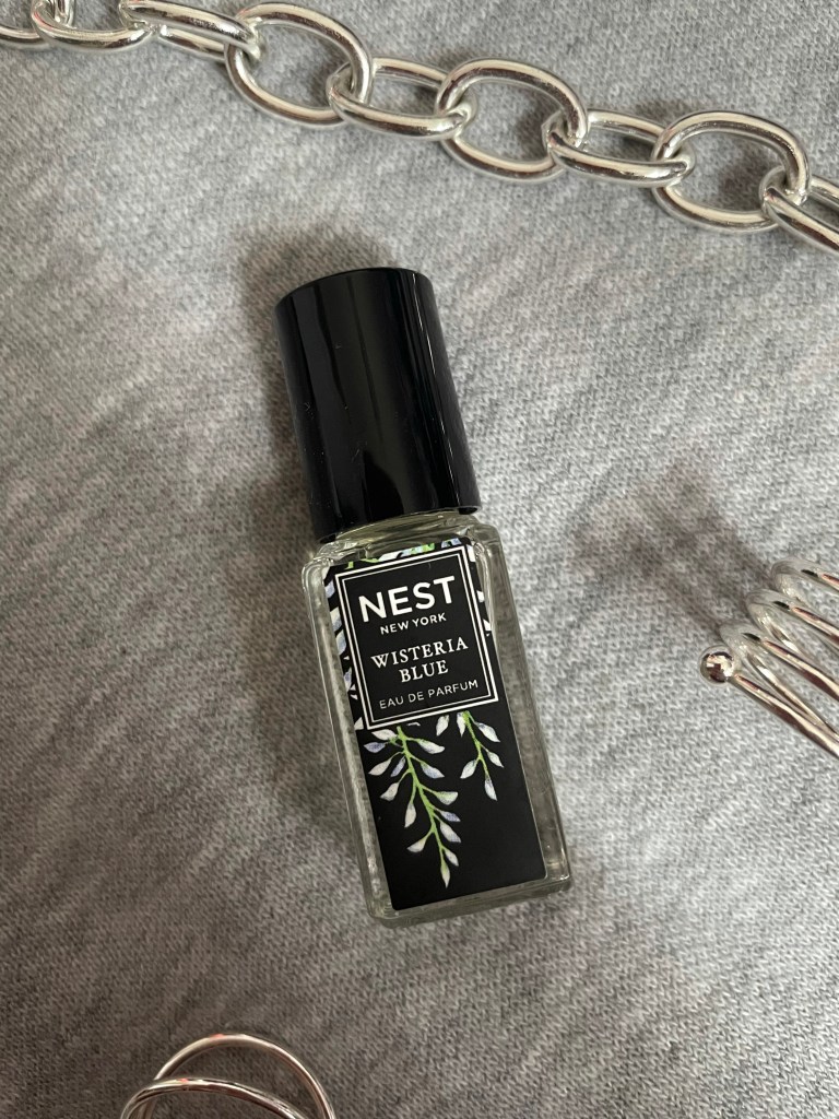A picture of the travel size fragrance Wisteria Blue from NEST New York Fragrances on a gray background surrounded by silver jewelry.