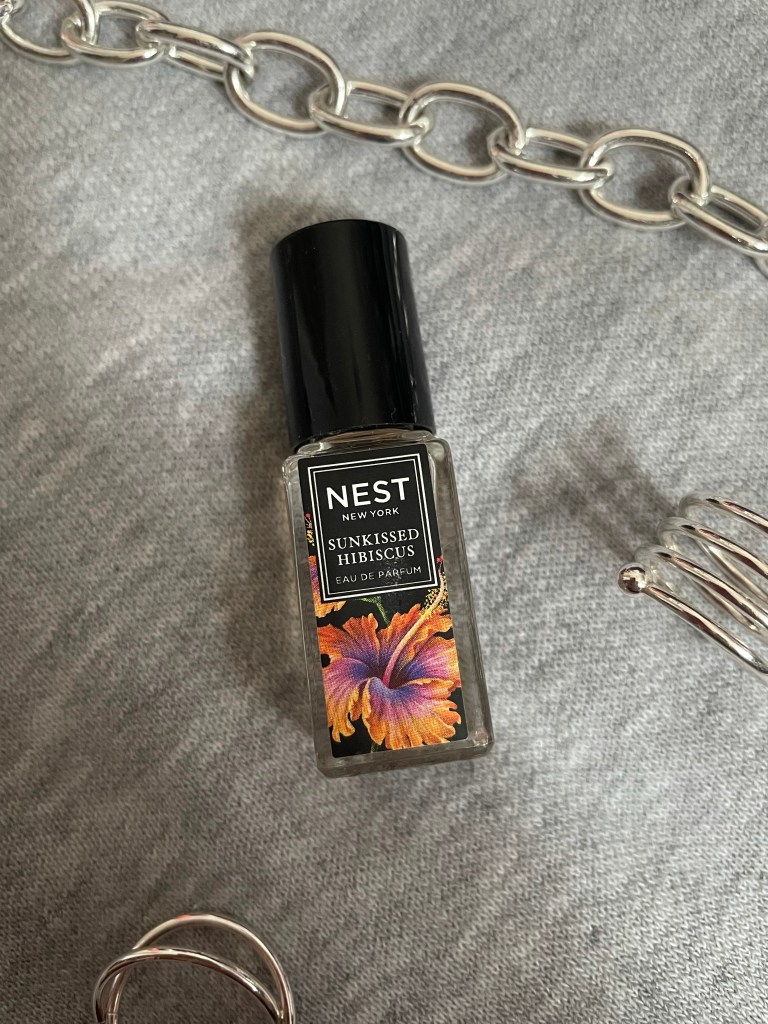 A picture of the travel size fragrance Sunkissed Hibiscus from NEST New York Fragrances on a gray background surrounded by silver jewelry.