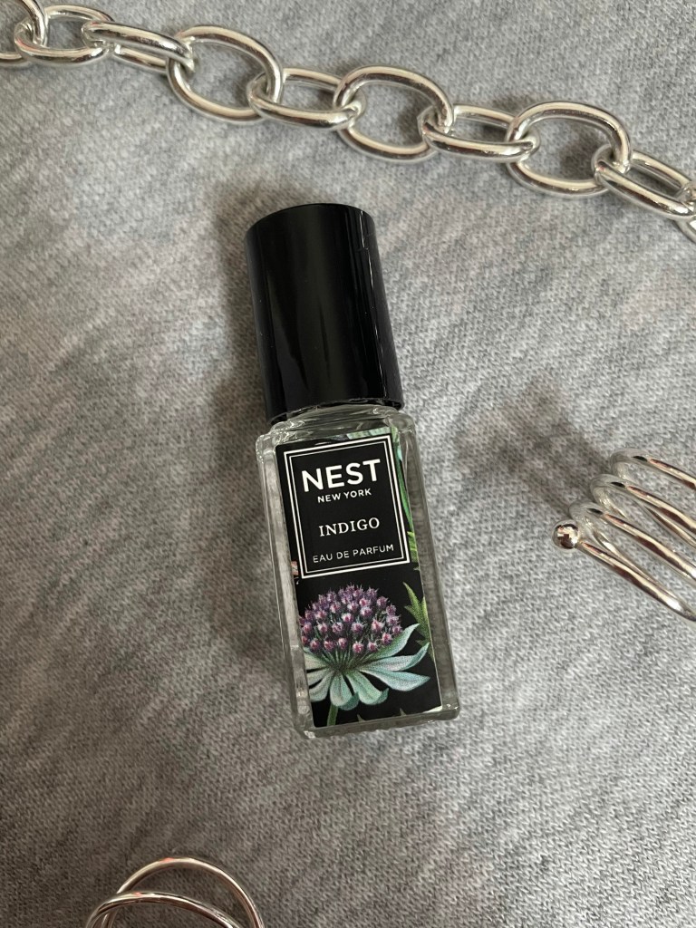 A picture of the travel size fragrance Indigo from NEST New York Fragrances on a gray background surrounded by silver jewelry.